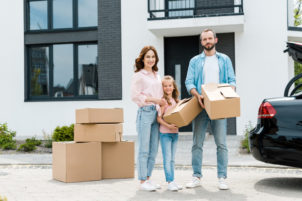 cheerful family standing with boxes while moving into modern house - Photo, Image