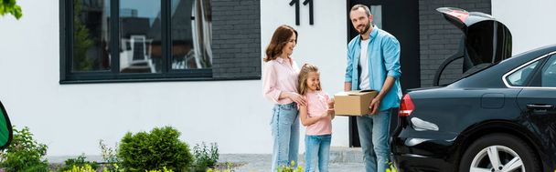 panoramische shot van vrolijke familie verhuizen naar modern huis terwijl staande in de buurt van auto  - Foto, afbeelding