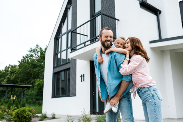 happy bearded man piggybacking daughter near cheerful wife and home  - Photo, Image