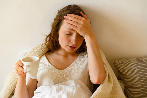 sick young woman covered with blanket holding cup of hot tea while lying in bed  - Photo, Image