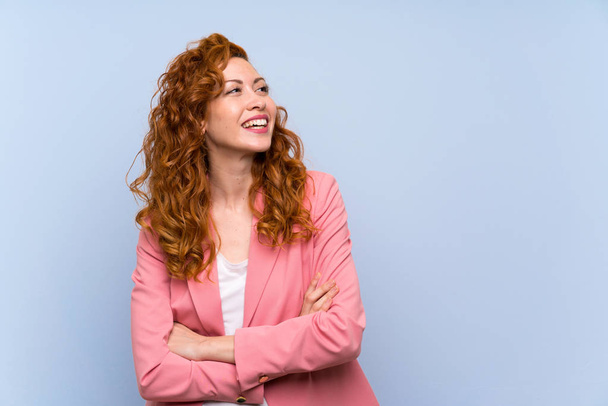 Redhead vrouw in pak over geïsoleerde blauwe muur gelukkig en glimlachend - Foto, afbeelding
