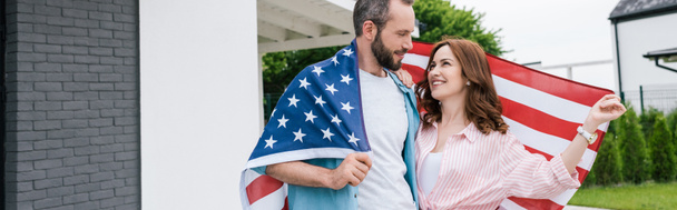 panoramiczny strzał szczęśliwego Brodatego mężczyzny stojącego z atrakcyjną kobietą i trzymając amerykańską flagę  - Zdjęcie, obraz