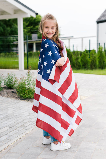 glückliches Kind lächelt, während es mit amerikanischer Flagge in der Nähe des Hauses steht  - Foto, Bild