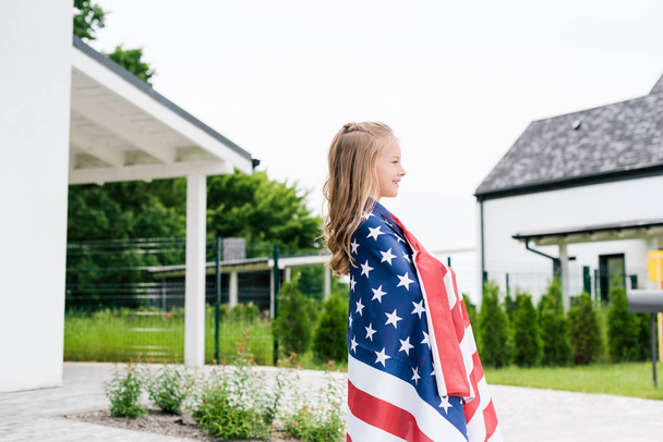 pohled ze strany šťastného kluka, který se usmívá, když stál s americkou vlajkou blízko domu  - Fotografie, Obrázek