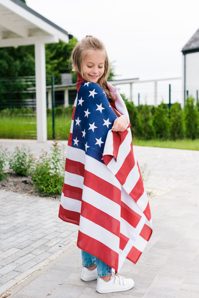 cheerful kid smiling while standing with american flag near house  - Φωτογραφία, εικόνα