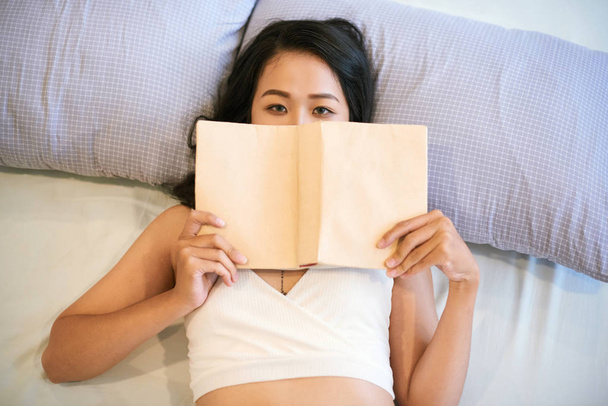 Portret azjatyckiej młodej kobiety leżącej na łóżku z książką patrząc na kamerę i będzie czytać - Zdjęcie, obraz