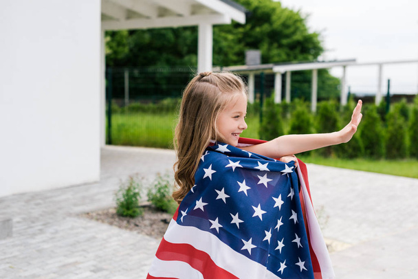 bambino felice sventolando mano mentre in piedi con bandiera americana vicino casa
  - Foto, immagini