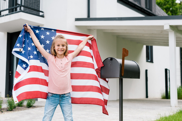 happy kid standing and holding american flag near mail box and house  - 写真・画像