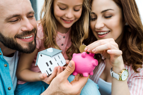 selective focus of happy woman putting golden coin in piggy bank near husband and daughter  - Photo, Image