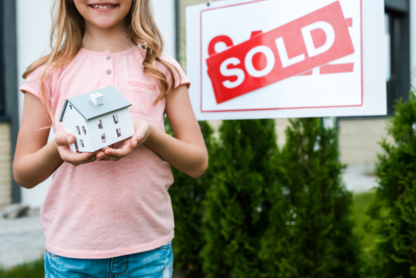 vista recortada de niño alegre sosteniendo modelo de casa de cartón cerca de la junta con cartas vendidas
  - Foto, Imagen