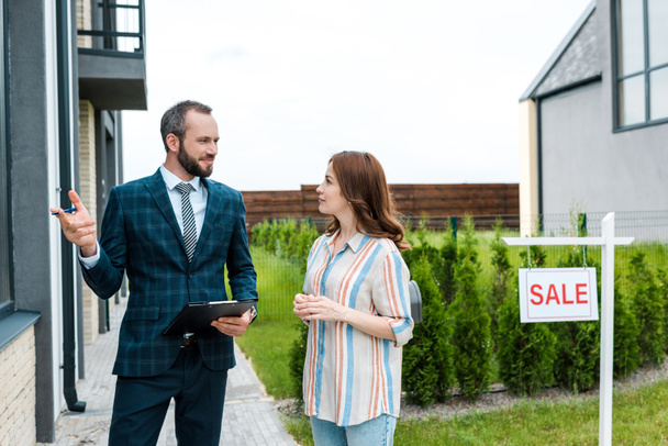 handsome bearded broker holding clipboard and gesturing near attractive woman and board with sale letters  - Photo, Image
