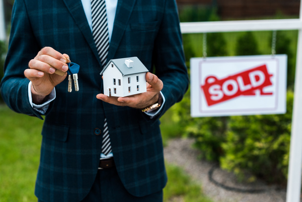 cropped view of realtor holding carton house model and keys near board with sold letters  - Фото, изображение
