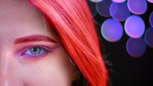 Closeup half face shoot of young attractive caucasian female with pink beautiful pink glitter makeup with coral red hair looking at camera with bokeh background - Footage, Video