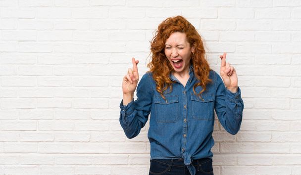 Redhead woman over white brick wall with fingers crossing - Photo, Image