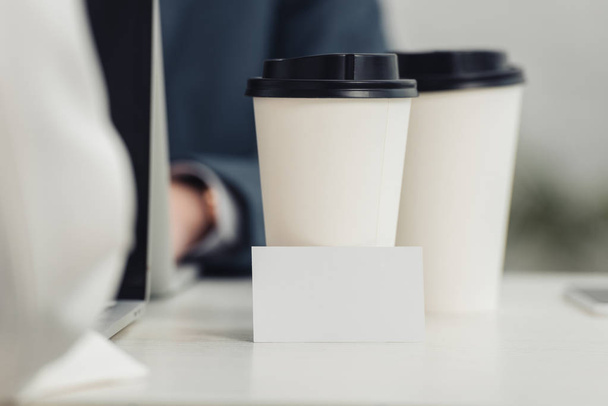 selective focus of blank business card and disposable cups on table near businesspeople using laptops - Фото, изображение