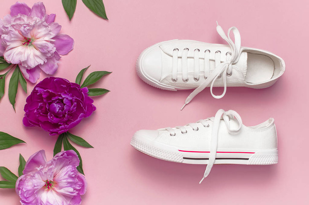White female fashion sneakers and pink purple flowers peonies on pink background. Flat lay, top view, copy space. Women's shoes. Stylish white sneakers. Fashion blog or magazine concept. - Photo, Image