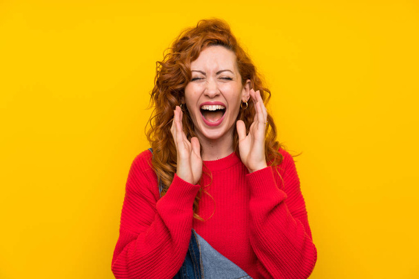 rothaarige Frau mit Overalls über isolierter gelber Wand lacht - Foto, Bild