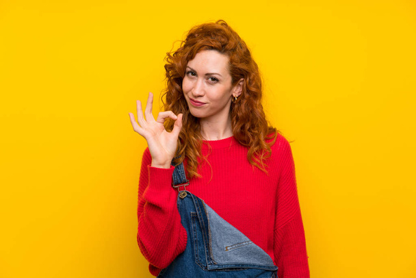 Femme rousse avec salopette sur mur jaune isolé montrant un signe ok avec les doigts
 - Photo, image
