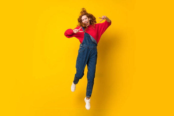 rothaarige Frau mit Overalls springt über isolierte gelbe Wand - Foto, Bild