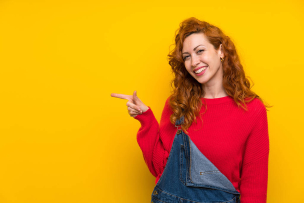 Redhead vrouw met overalls over geïsoleerde gele muur wijzende vinger aan de zijkant - Foto, afbeelding