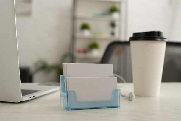 держатель карт с белыми пустыми визитными карточками возле бумажной чашки и ноутбука на рабочем столе
 - Фото, изображение