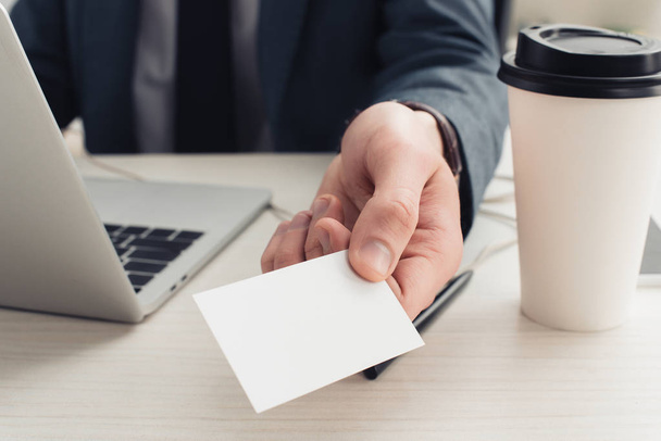 vista cortada de empresário segurando cartão de visita vazio enquanto sentado perto de laptop e café para ir
 - Foto, Imagem