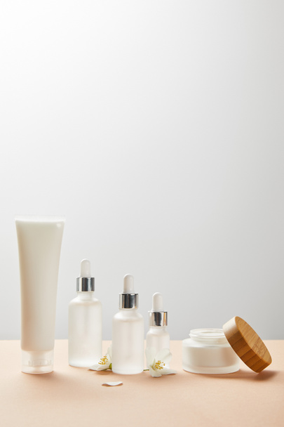 tubo de crema con crema, botellas de vidrio cosmético con suero, frasco abierto con tapa de madera y pocas flores de jazmín en beige
  - Foto, Imagen