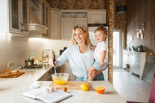 Young mother and her daughter mixing fruits in a bowl. Cute woman and little girl cooking on the kitchen, cake preparation. Happy family prepares sweet dessert, ingredients on the table - Photo, Image