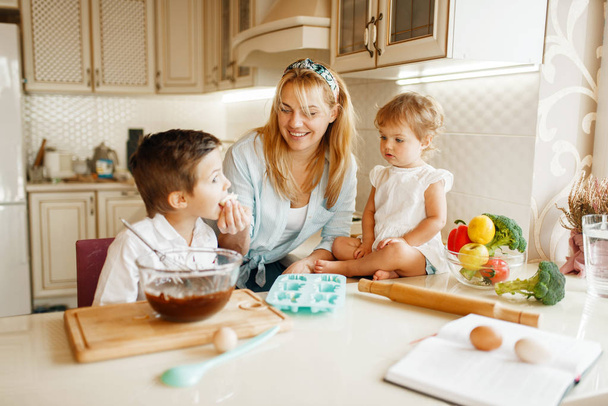 Young mother and her kids tastes fresh pastry with melted chocolate. Woman and little children cooking on the kitchen, cake preparation. Happy family eats sweet dessert - Foto, immagini