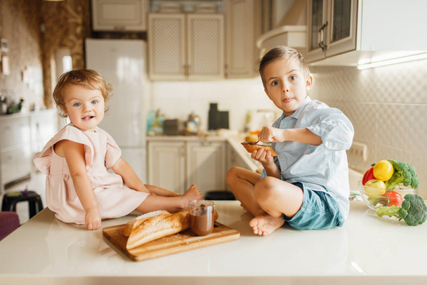 Dos niños untan chocolate derretido en pan, sabrosos sándwiches. Lindo chico y chica cocinando en la cocina. Los niños felices preparan y saborean postre dulce en el mostrador
 - Foto, Imagen