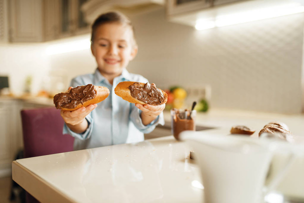 Το νεαρό αγόρι δείχνει σάντουιτς με φυσική λιωμένη σοκολάτα. Χαριτωμένο αρσενικό παιδί που μαγειρεύει στην κουζίνα. Ευτυχισμένο παιδί προετοιμάζει και έχει γεύση γλυκό επιδόρπιο στο τραπέζι - Φωτογραφία, εικόνα
