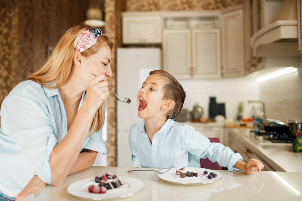 Young mother with her kid tasting chocolate pastry. Cute woman and little boy cooking on the kitchen. Happy family tasting sweet dessert at the counter and having fun - Foto, afbeelding
