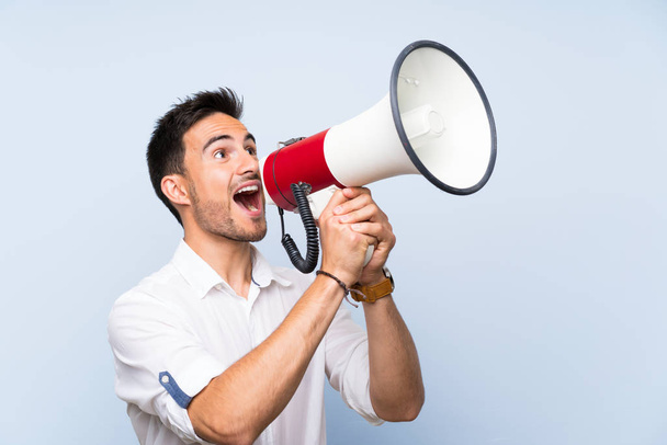 Handsome young man over isolated blue background shouting through a megaphone - Foto, Imagen