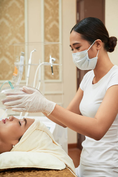 Joven cosmetóloga en uniforme haciendo spa procedimiento facial para la cara de su cliente en salón de belleza
 - Foto, Imagen