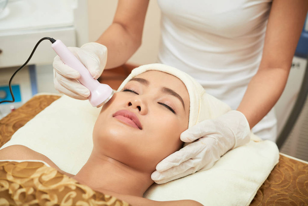Asiatico bella donna sdraiata con gli occhi chiusi mentre cosmetologo facendo ultrasuoni procedura per il suo viso in moderno centro benessere salone
 - Foto, immagini