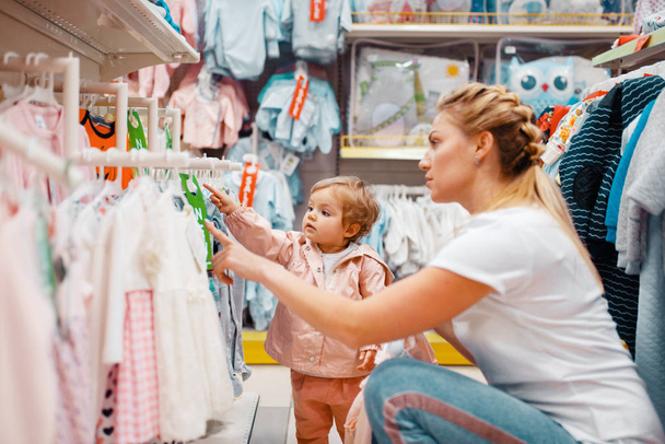 Мама со своей маленькой девочкой выбирает одежду в детском магазине. Мама и ребенок вместе покупают платье в супермаркете
 - Фото, изображение