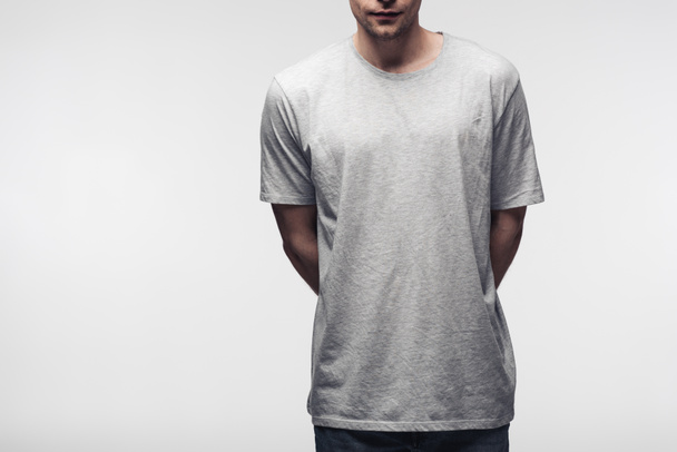 gedeeltelijke weergave van de mens in grijs t-shirt houden handen achter rug geïsoleerd op grijs, menselijke emotie en expressie concept - Foto, afbeelding