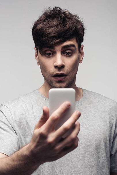 verrast jonge man kijken naar smartphone geïsoleerd op grijs - Foto, afbeelding