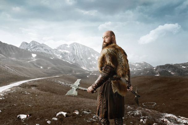 Viikinki kirveet käsissä pukeutunut perinteisiin pohjoismaisiin vaatteisiin seisoo kallioiset vuoret. Skandinaavinen muinainen soturi
 - Valokuva, kuva