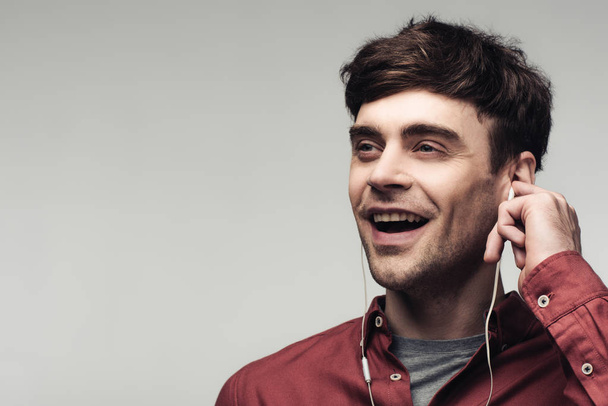 cheerful young man looking away while listening music in earphones isolated on grey - Foto, imagen