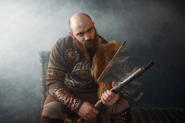 Barbudo viking com machado vestido com roupas nórdicas tradicionais sentado na cadeira, imagem bárbara. Guerreiro antigo em fumaça no fundo escuro
 - Foto, Imagem