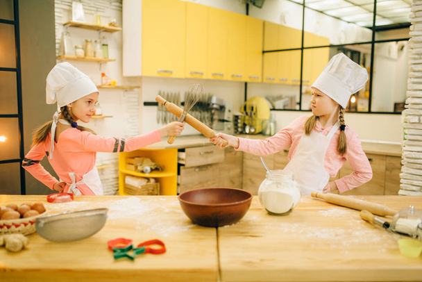 Dos chicas cocinan en peleas de gorras en la cocina. Niños cocinando pastelería, pequeños chefs sostiene rodillo y batidor para azotar
 - Foto, imagen