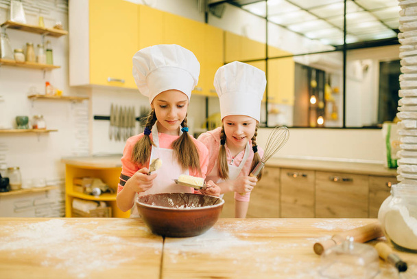 Дві маленькі дівчата готують в шапках додають масло в миску, готують печиво на кухні. Дитяче кулінарне тісто, дитячі кухарі роблять тісто, дитячий торт
 - Фото, зображення
