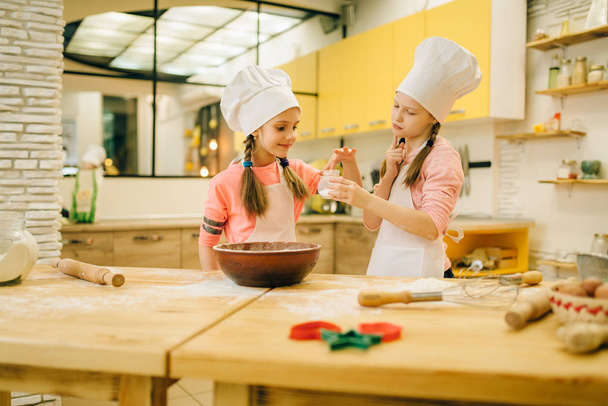 キャップで調理する2人の小さな女の子は甘いバニラパウダー、キッチンでクッキーの準備を味わいます。子供たちはペストリーを調理し、ケーキを準備する子供のシェフ - 写真・画像