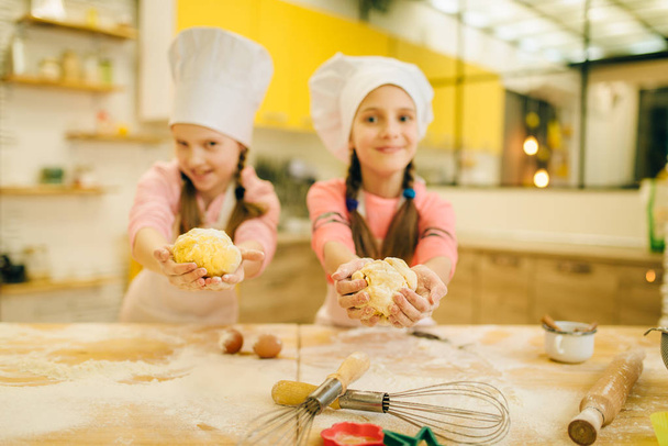 Kaksi hymyilevää pikkutyttöä kokkaa korkeissa näyttää taikinapalloja, evästeitä keittiössä. Lapset ruoanlaitto leivonnaiset, lapset kokit valmistelevat kakkua
 - Valokuva, kuva