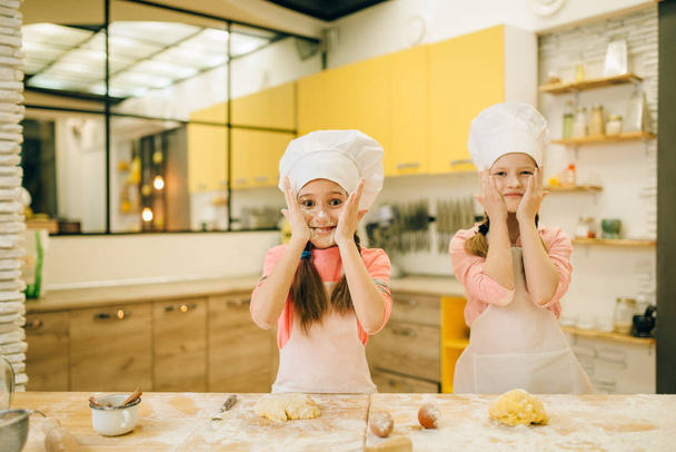 二人の小さな女の子のシェフは、キッチンで小麦粉、クッキーの準備で自分の顔を塗ります。子供たちはペストリーを調理し、楽しみを持って、子供たちはケーキを準備します - 写真・画像