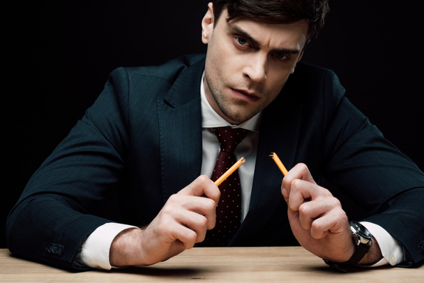 serious businessman holding broken pencil and looking at camera isolated on black - Фото, изображение