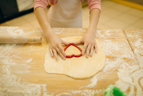 Little girl cook in apron makes cookies in the shape of a heart on the kitchen. Kid cooking pastry, child chef preparing cake - Photo, Image