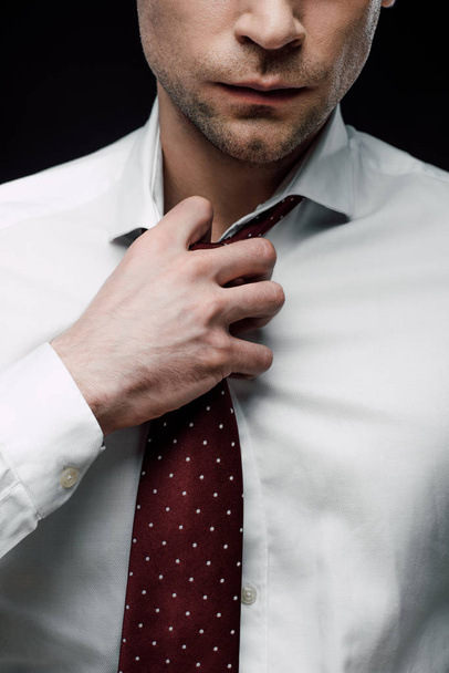 partial view of serious businessman touching tie isolated on black - Valokuva, kuva