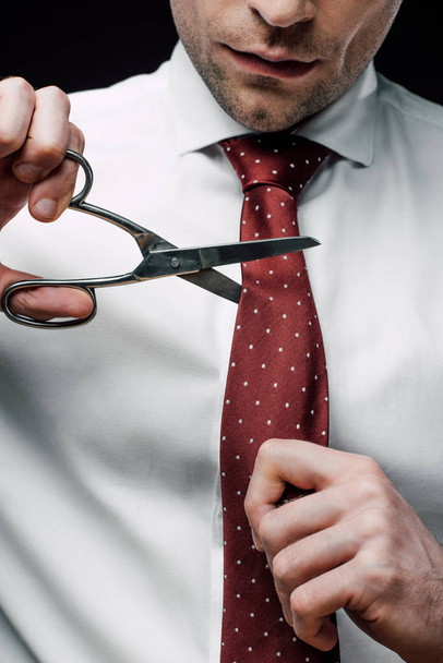 cropped view of displeased businessman cutting tie with scissors isolated on black - Photo, Image